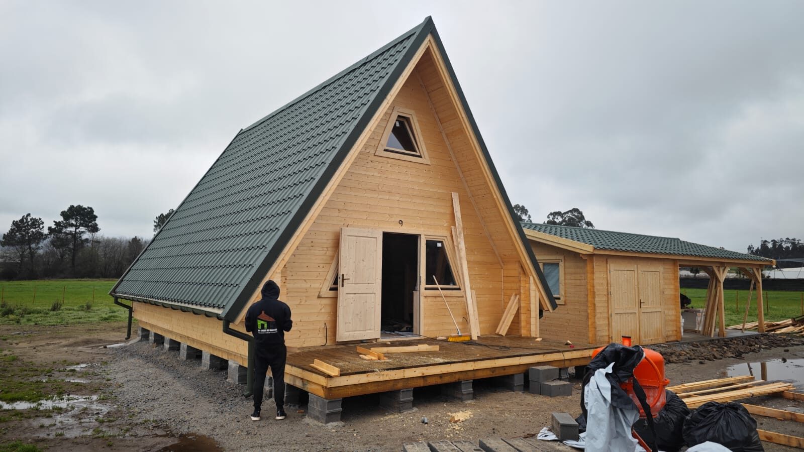 Casa A-Frame fechada com telhado verde, portas abertas e deck, ao lado do anexo de garagem