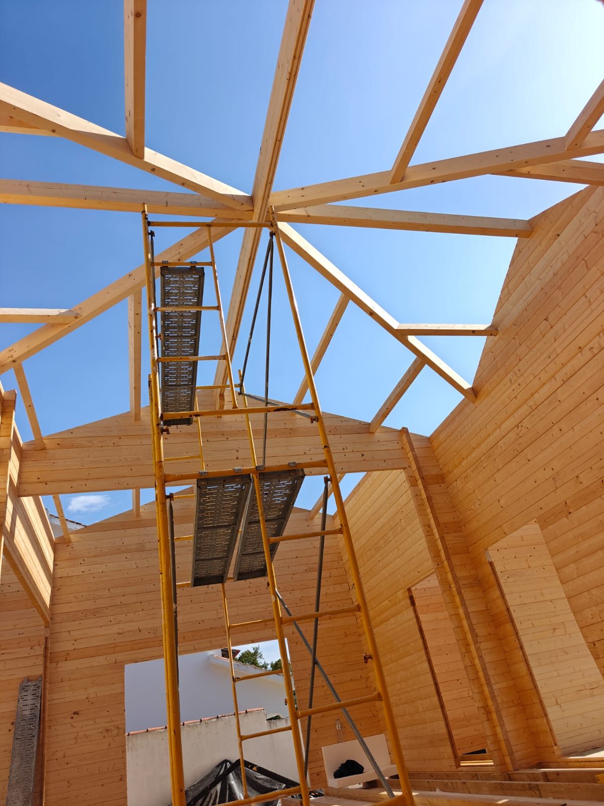 Interior da Villa T4 com a estrutura de vigas da cobertura contra o céu azul e o andaime da equipa de montagem